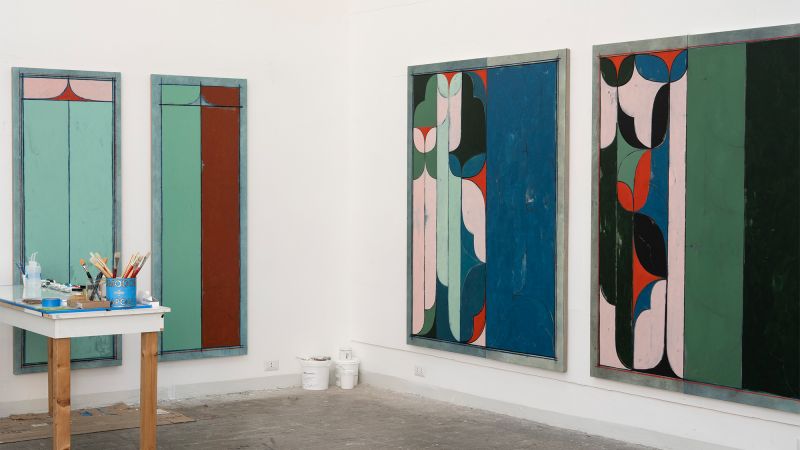 Color photo of three colorful abstract paintings of the same size hanging on two adjacent walls, with a work table in front of one
