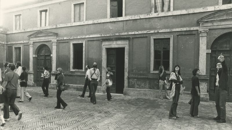 black and white photo of various people standing or walking on a tiled sidewalk in Siena, with a building behind them