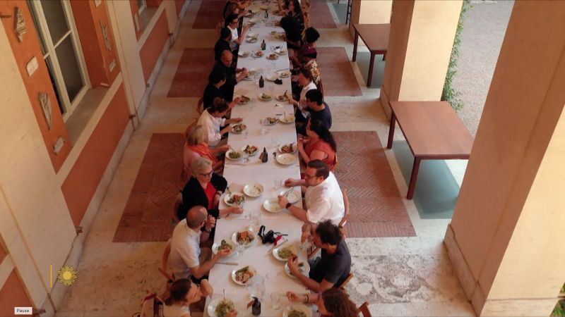 An aerial view of a long lunch table with people seating at it, conversing and eating