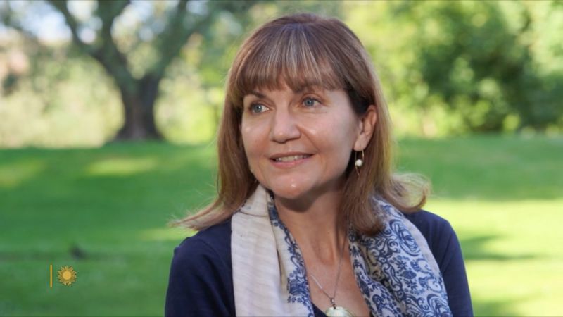 A video still of a light skinned woman sitting in an outdoor garden location and looking at an offscreen interviewer to her right
