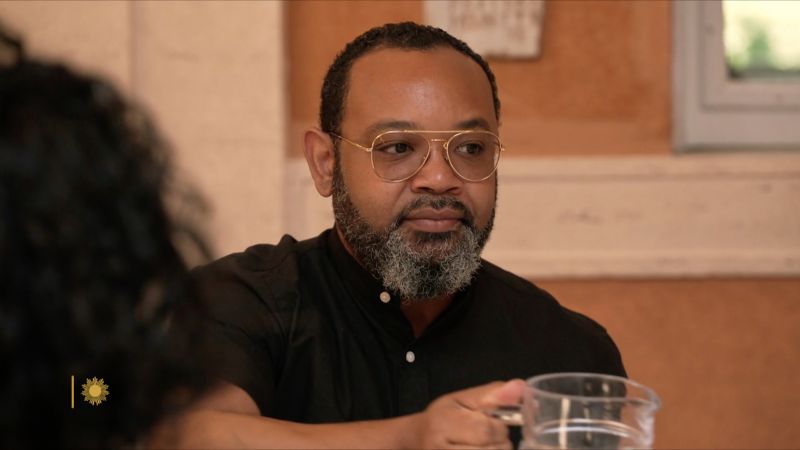 Color video still of a dark skinned, gray beared man seated at a lunch table and passing a water jug