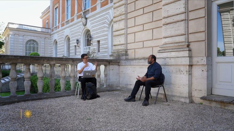 Color video still of an Asian and black man seated in chairs and talking to each other; they are on the porch of the Academy building next to a studio
