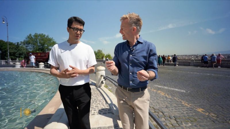 An Asian man and a light skinned man talk to each other and walk alongside the Fontanone in Rome on a clear, sunny day