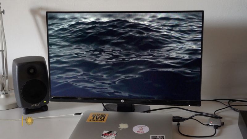 Color video still of a computer monitor showing ocean waves; also on the desk are a laptop and one audio speaker