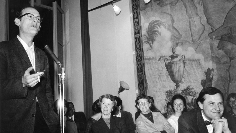 Black and white photo of a white man speaking into a microphone as seated women and men watch him