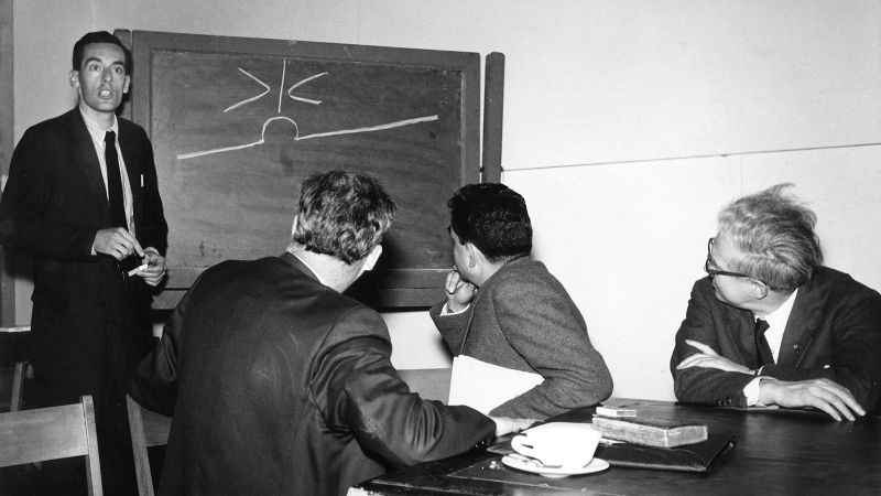 Black and white photograph of three white men sitting at a table looking at a fourth man standing at a chalkboard