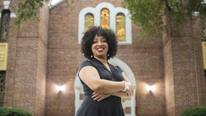 Color photograph of a dark skinned woman standing in front of a brick chapel; she wears a black dress, crosses her arms, and smiles at the camera