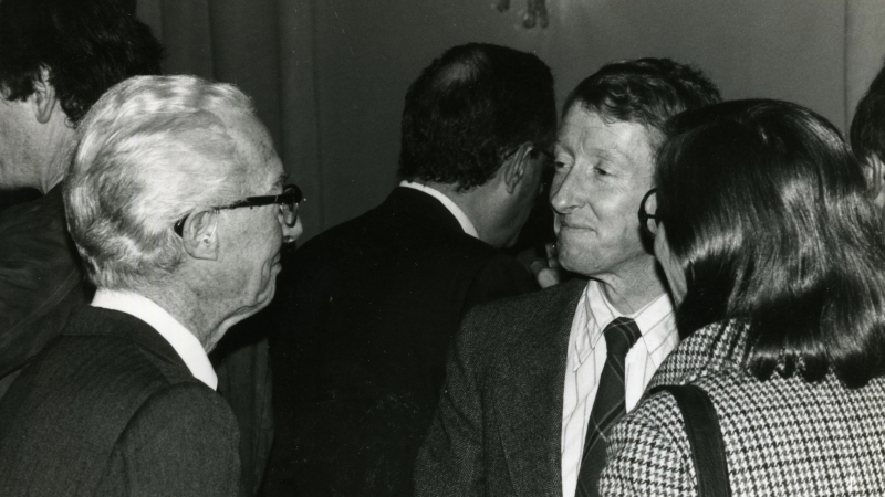 A black and white photo in which a man in a tie speaks with another man