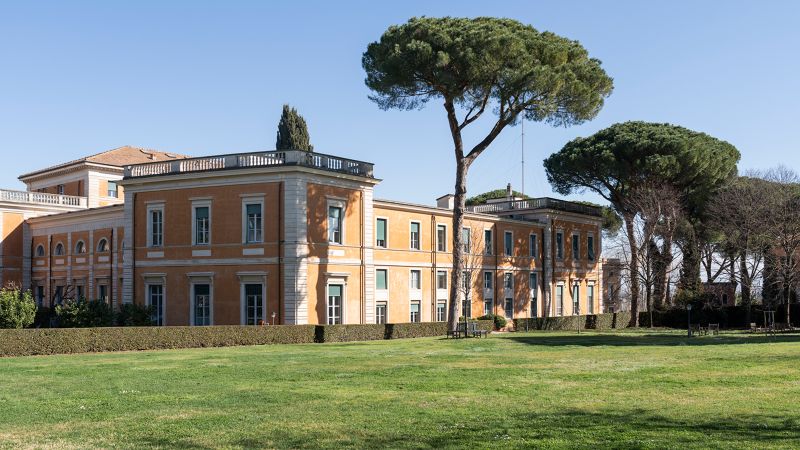 Color photo of an Italian villa influenced building, a grassy lawn, and umbrella pines on a sunny day