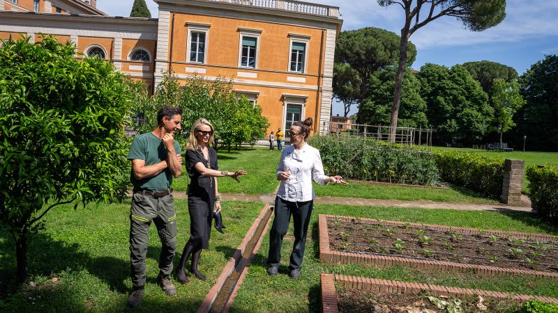 Color photo of two women and one man looking at garden beds, with a building and trees behind them
