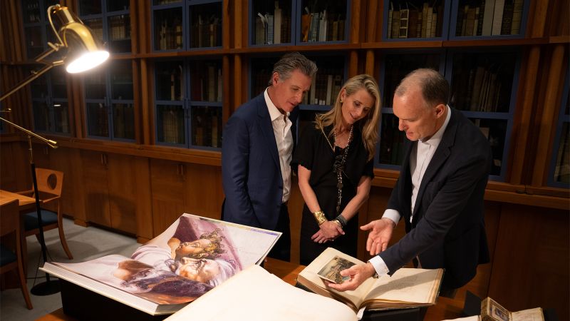 Color photograph of two light skinned men and one light skinned woman looking at rare books in a library