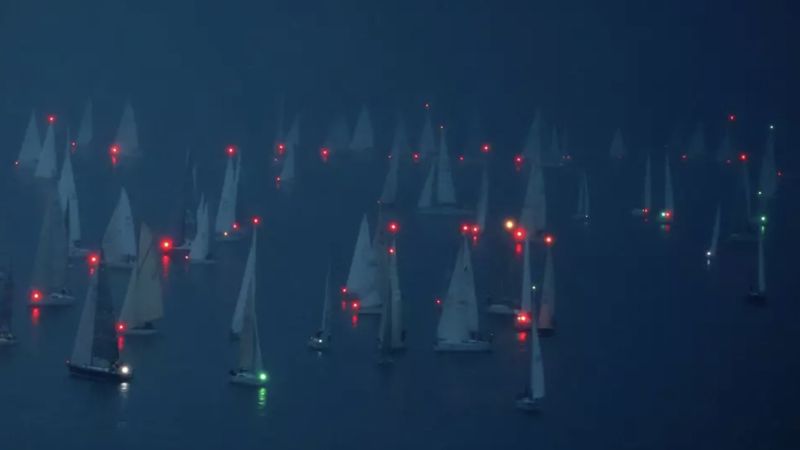 Color photograph of what looks like many sailboats in water, each with a red light at its mast
