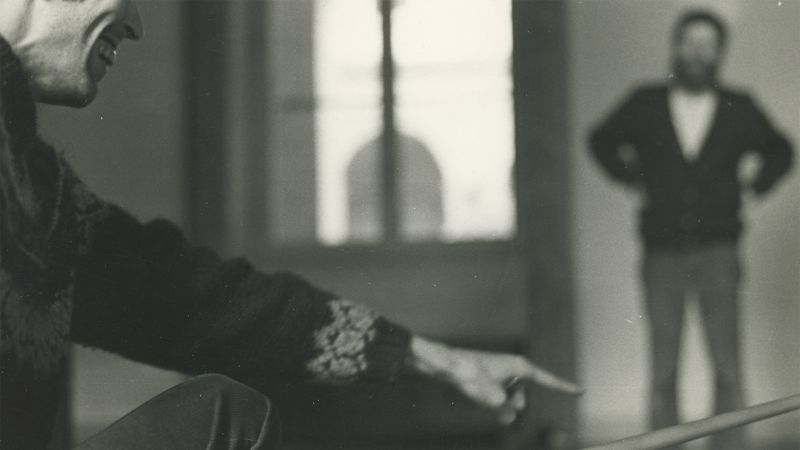 Black and white photo of a light skinned man holding a cue stick; away from him and out of focus is a standing light skinned man