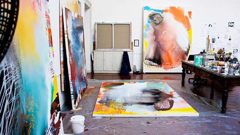 An artist's studio with abstract paintings in progress that are leaning against walls or laying on the floor, as well as a table covered with paint cans, brushes, and other supplies