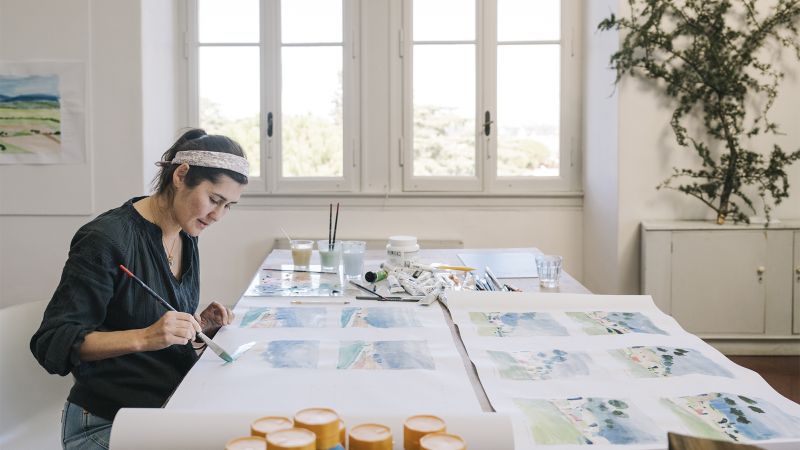 Color photo of a light skinned woman in an artist studio, sitting at a work table and painting sheets of paper