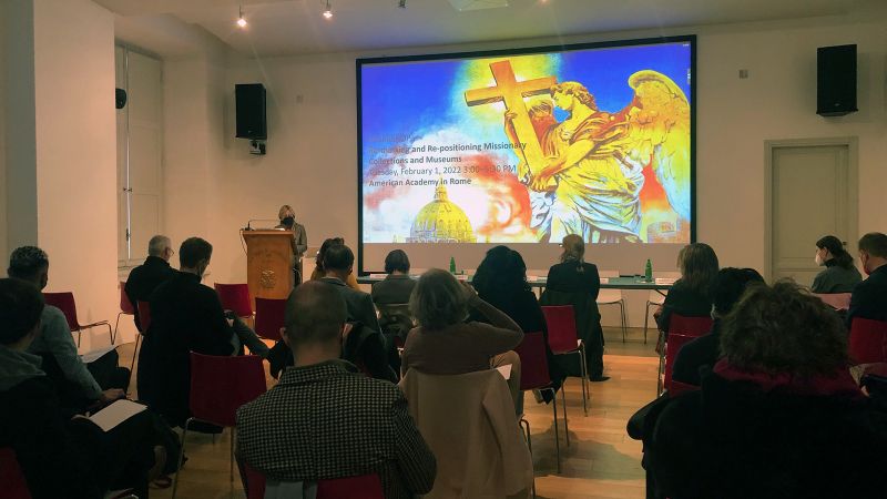 Color photograph of a lecture room seen from the rear, showing people sitting in chairs looking at a speaker at the podium or a digital image projected on the screen