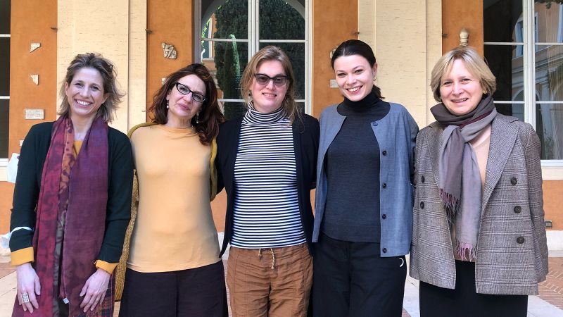 Color photograph of five light skinned women of varying ages standing in a row in an outdoor location, looking at the camera and smiling