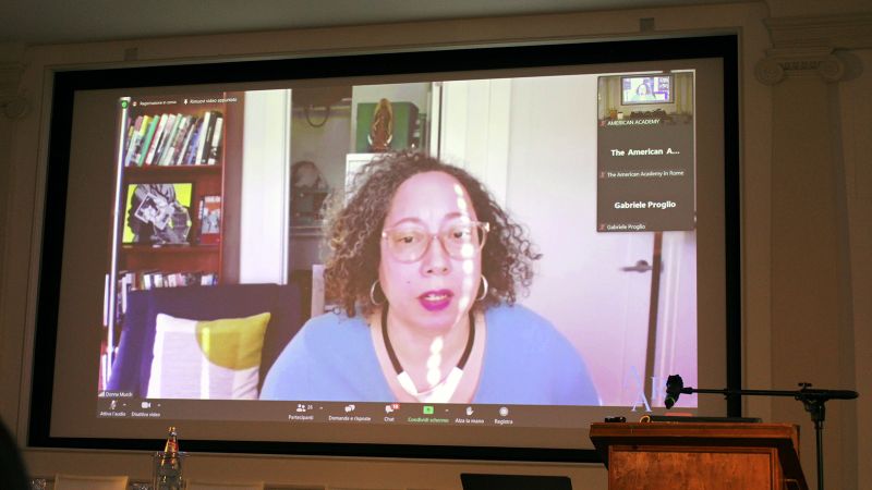 Color photograph of the projection screen of a lecture hall showing a medium skinned man, wearing glasses and sitting inside a home or office, giving a lecture remotely