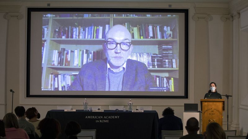 Color photograph of the projection screen of a lecture hall showing a light skinned man, wearing glasses and sitting in front of a full bookshelf, giving a lecture remotely
