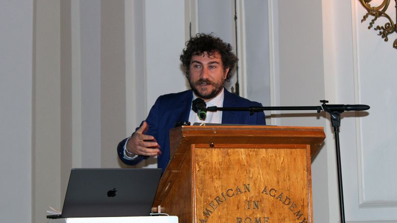 Color photograph of a light skinned man with dark curly hair and a beard speaking at a podium in an indoor lecture room; he gestures with one arm
