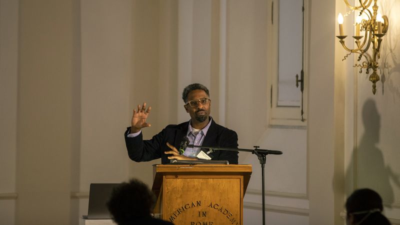 Color photograph of a dark skinned man speaking at a podium in a lecture hall, holding up one arm to emphasize a point