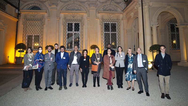 Color photograph of a group of 14 men and women of varying ages and ethnicities standing outdoors at night on a gravel lawn in front of an Italian villa