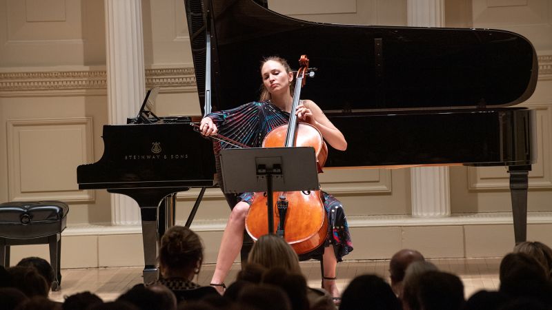 Color photo of a light skinned woman playing the cello onstage with a grand piano behind her