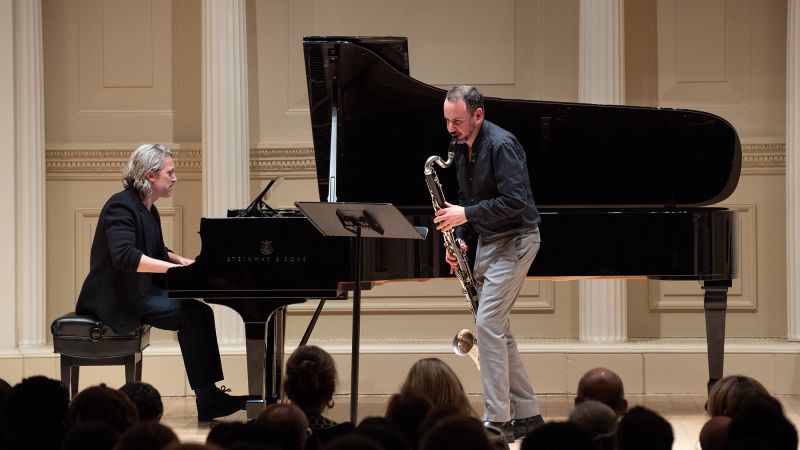 Color photo of two musicians on stage, one seated at a piano, another standing and holding a bass clarinet