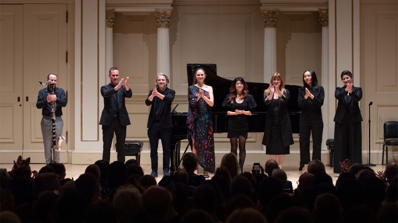 Color photo of eight musicians standing onstage after a concert, applauding the crowd and each other