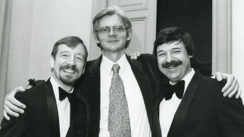 Black and white photograph of the heads and torsos of three light skinned men posing for the camera: a tall man in the center wears a jacket and tie; the two shorter men flanking him wear jackets and bowties and sport mustaches