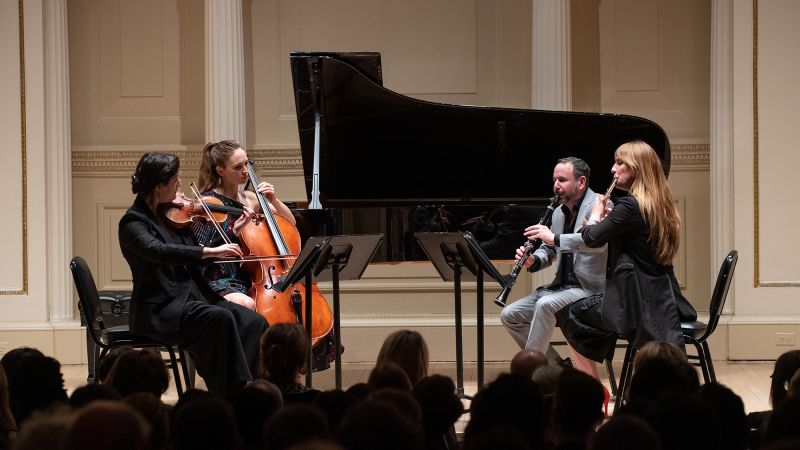 Color photo of four seated musicians onstage, with music stands in front of them and a grand piano and curtained walls behind them