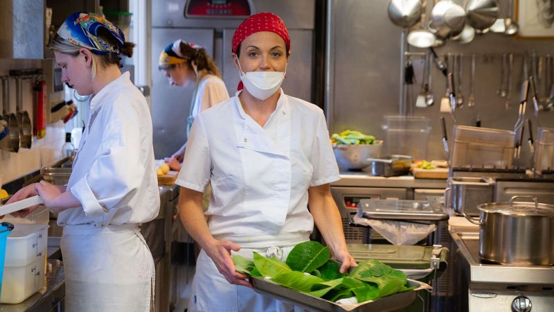 Color photograph of three light skinned woman in a kitchen cutting and transporting vegetables
