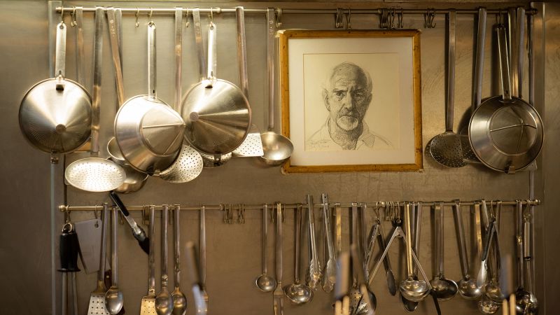 Color photograph of a commercial kitchen wall with two rows of steel utensils hanging from the wall, alongside a drawn portrait of a man's head