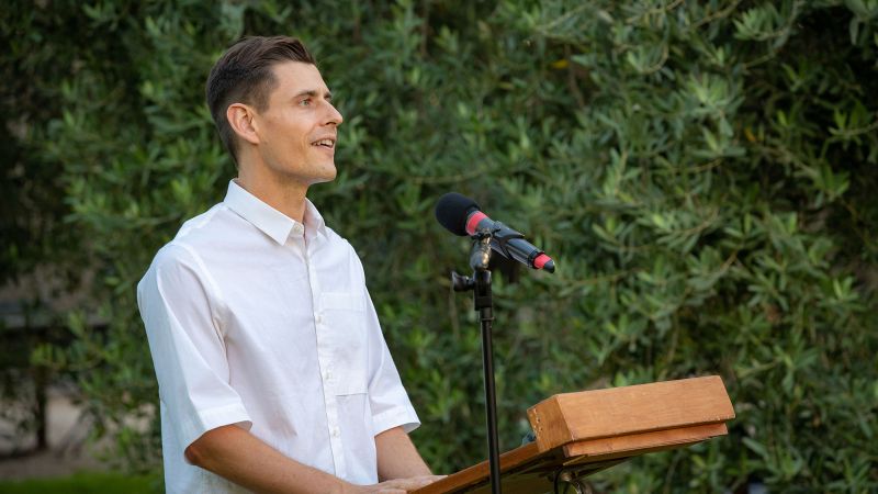 Color photograph of a light skinned man wearing a white shirt and speaking from a podium in a garden location