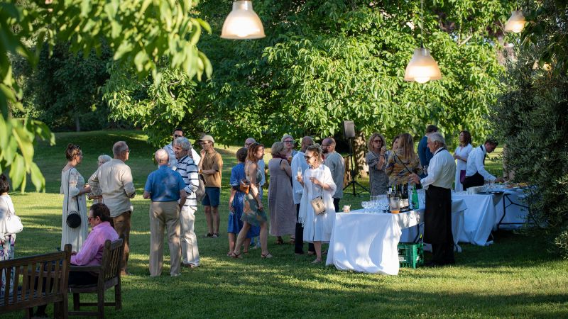 Color photograph of a couple dozen people standing in a garden near a catering table