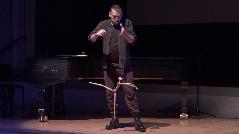 Color photograph of a light skinned man operating a puppet by its strings