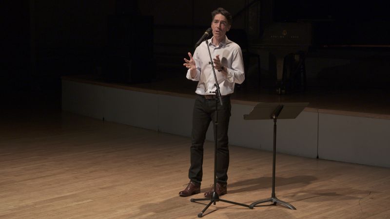 Color photograph of a light skinned man in a white shirt and dark pants speaking and gesturing to an unseen audience