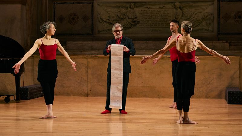 Color photo of three dancers surrounding a man reading from a paper scroll he is holding
