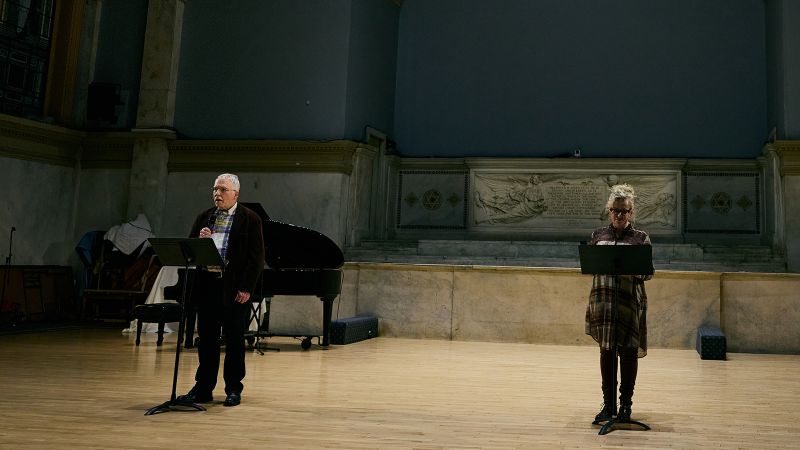 Color photo of a man and woman on a wood-floored performance stage; each has a microphone and a music stand with materials from which they read