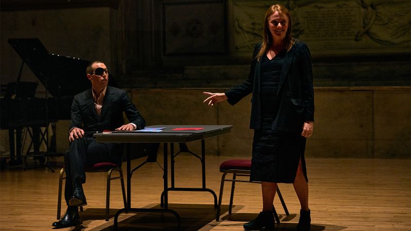 Color photograph of a play performance: a man sits in a chair at a table and a woman stands nearby