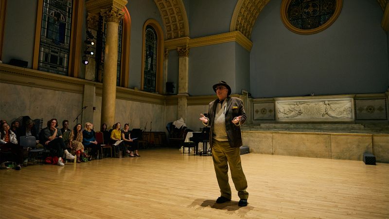 A man standing in a ballroom performance space speaking and gesturing toward the audience