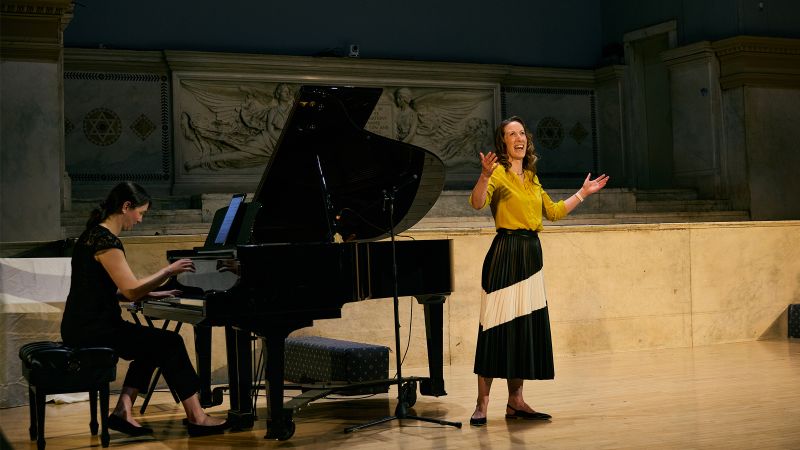 Color photo of a musical performance with one woman seated at a piano and another woman standing and singing