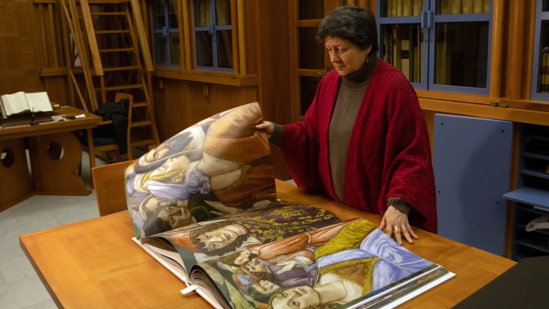 Color photograph of a light skinned woman leafing through a large book of color reproductions, which rests on a table is a room lined with full bookshelves