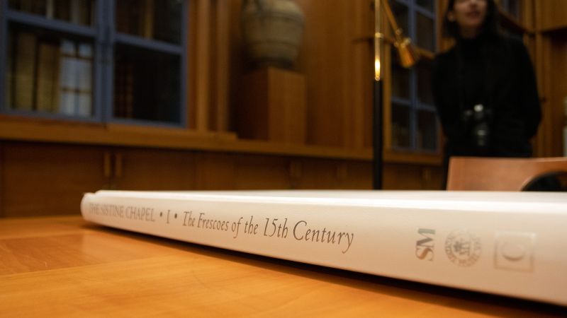 A color photograph depicting the white-clothed spine of a large book laying on a wooden library table