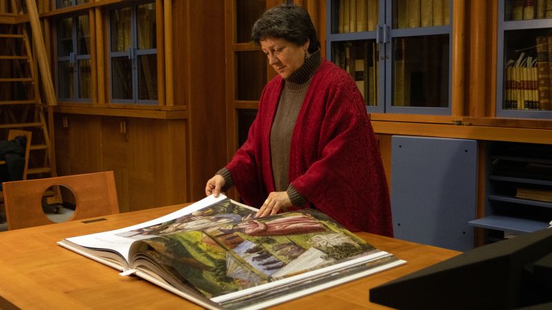 Color photograph of a light skinned woman leafing through a large book of color reproductions, which rests on a table is a room lined with full bookshelves