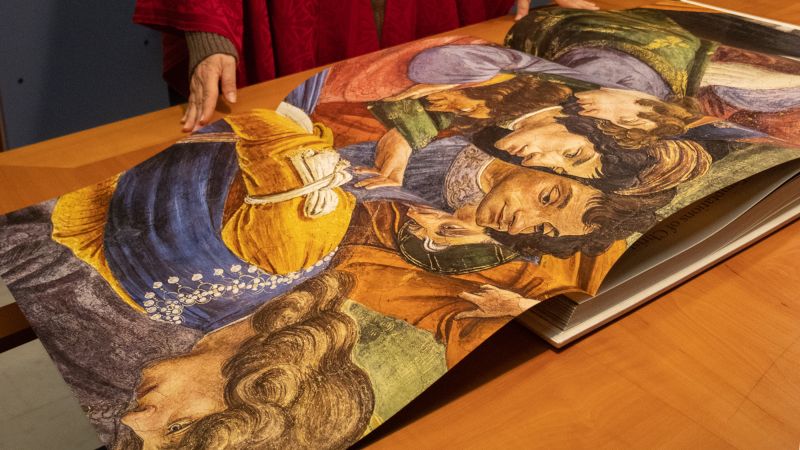 Color photograph of a large book opened to a gatefold spread depicting a detailed view of a Sistine Chapel fresco