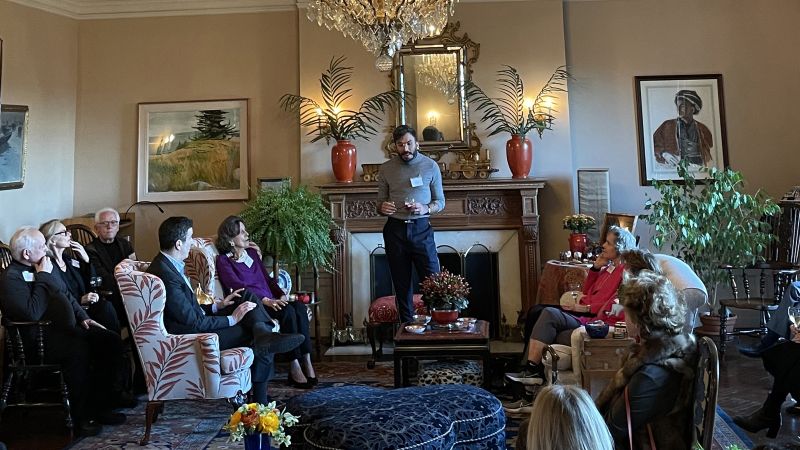 Color photograph of about twelve adults gathered in an ornately decorated living room; one man speaks as the others listen