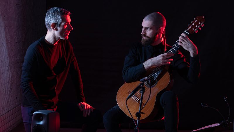 Two light skinned men talk to each other; one sits on a stage and holds an acoustic guitar; the other stands to his right
