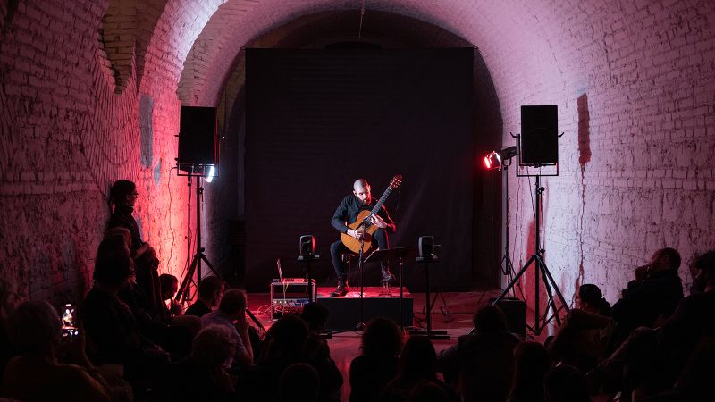 Color photograph of people watching a man play acoustic guitar on a small stage in a barrel-vaulted room