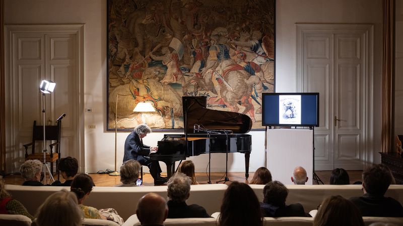 Color photograph of a man playing piano in a salon with a large tapestry on the wall behind the instrument